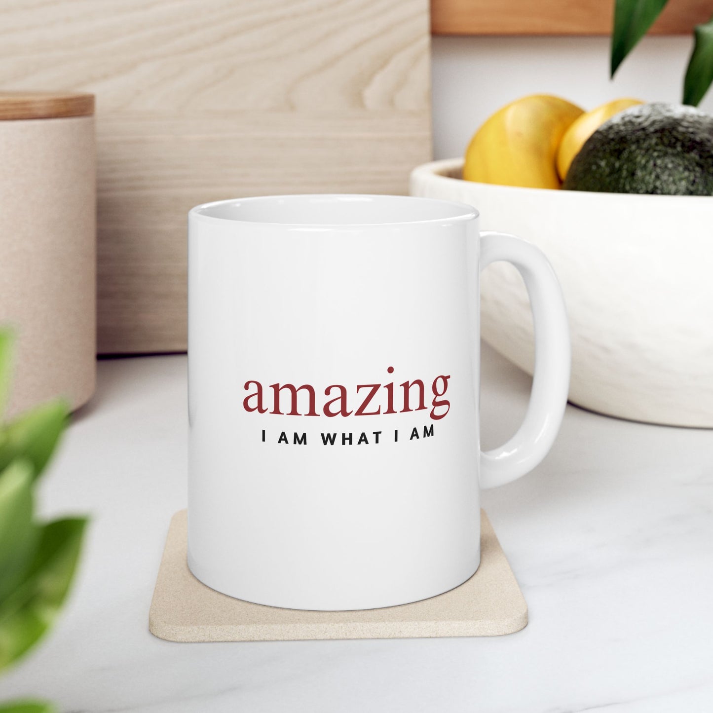 White mug with 'amazing I AM WHAT I AM' text on a coaster, surrounded by plants and a bowl of fruit.