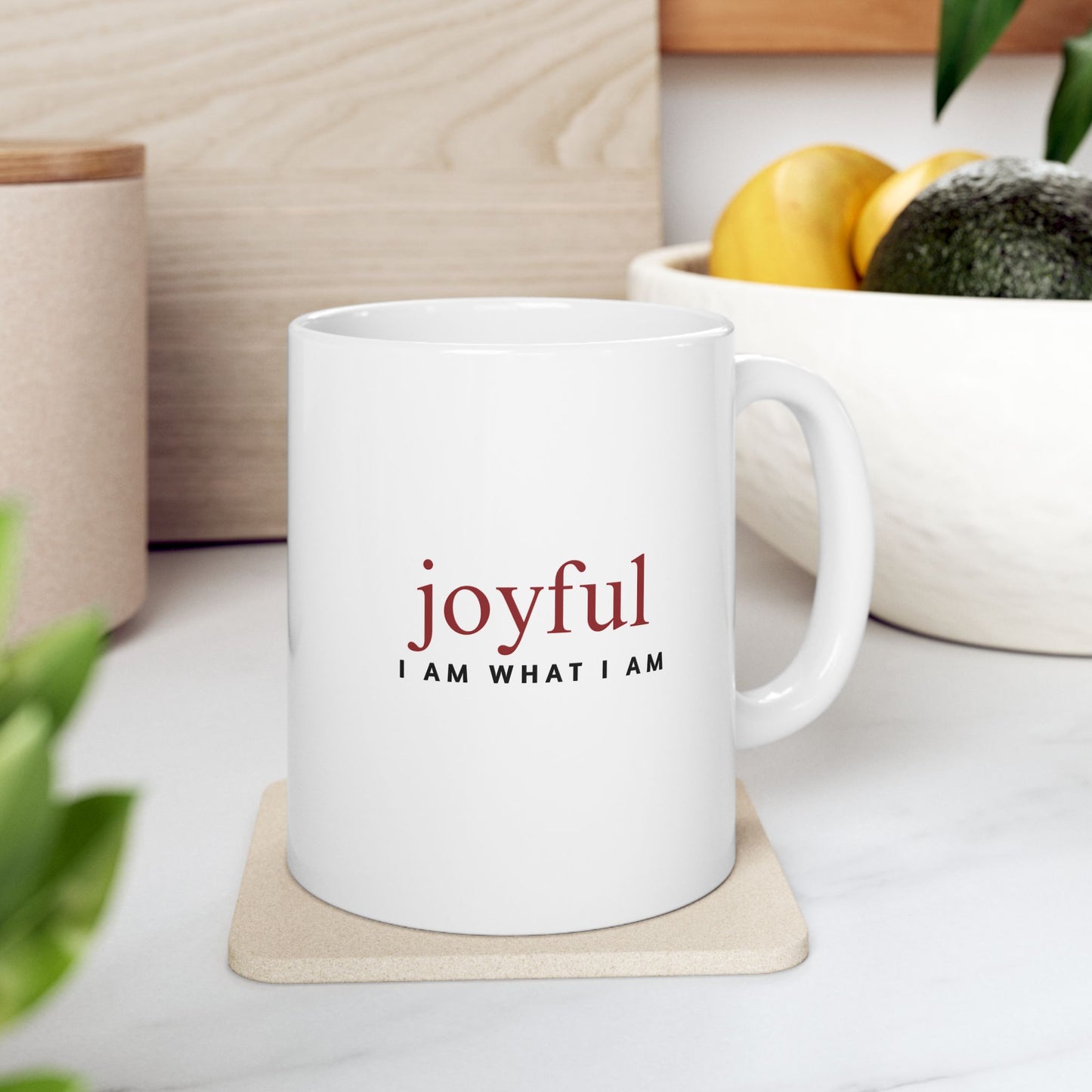 White mug with 'joyful I AM WHAT I AM' text on a coaster, surrounded by plants and a bowl of fruit.