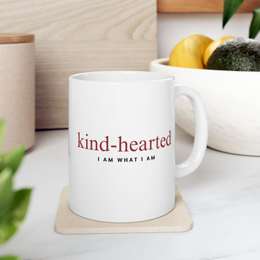White mug with 'kind-hearted I AM WHAT I AM' text on a coaster, surrounded by plants and a bowl of fruit.